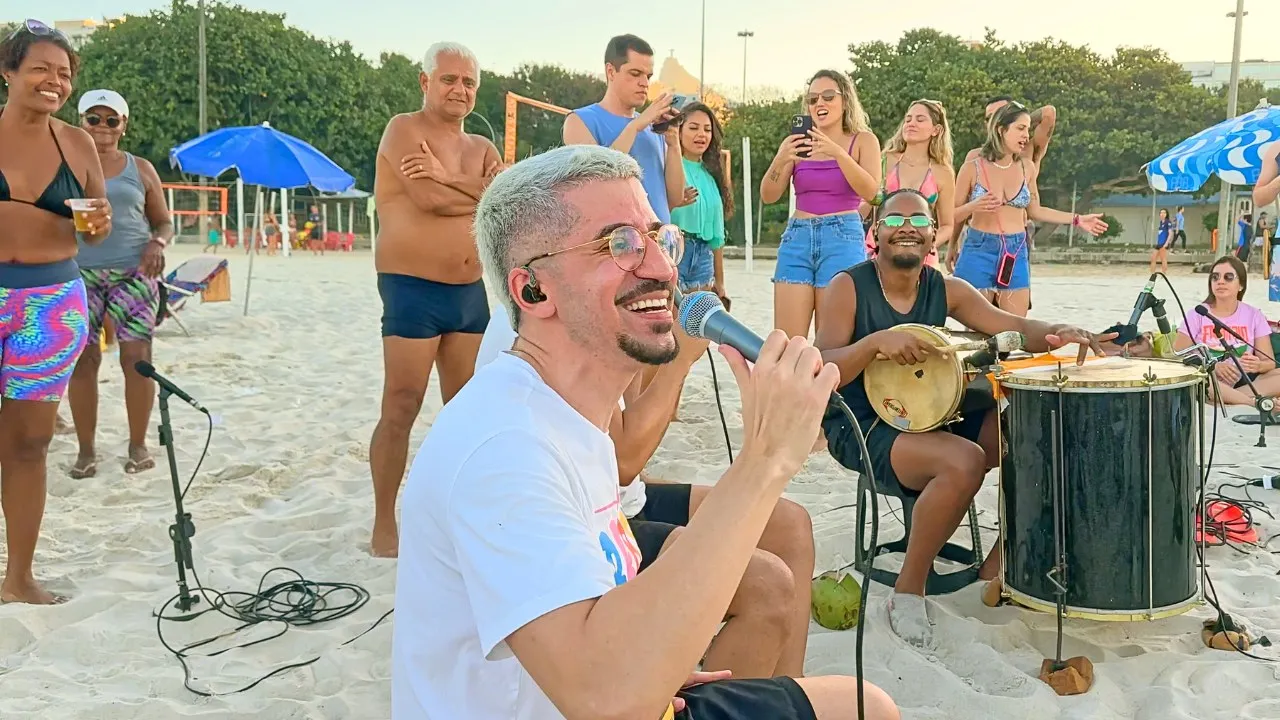 Leleco sorri cantando ao microfone, cercado pela roda, no fim de tarde